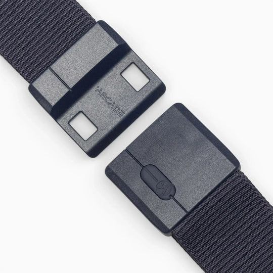 A close-up of the Aero Mag belt by Arcade Belts shows a black fabric strap with a durable buckle, partially unfastened, displaying the word "ARCADE" on one side. The buckle is equipped with two square holes, a locking mechanism, and features a tension lock for secure fastening.