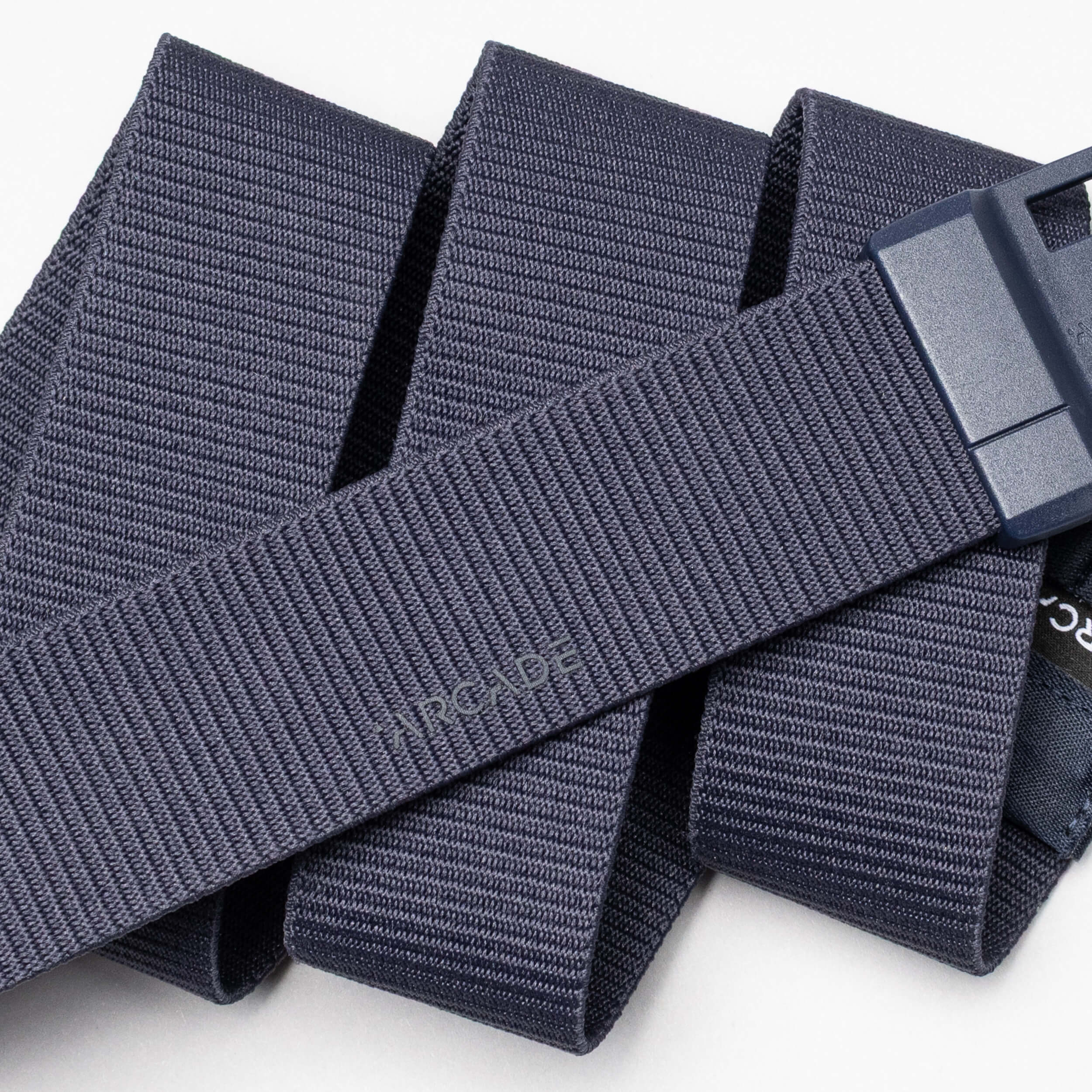 A close-up image of the Aero belt from Arcade Belts, featuring navy blue textured fabric and a dark plastic buckle, shown in overlapping folds on a white surface. "Arcade" is subtly printed on the belt.