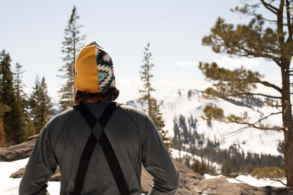 A person wearing a patterned yellow beanie and suspenders with a sleek plastic buckle stands outdoors, facing snow-covered mountains and pine trees under a clear sky.