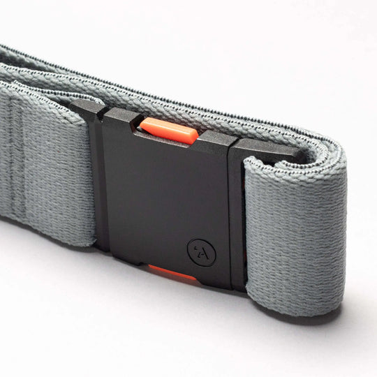 A close-up of the Arcade Belt Co. performance stretch belt by Arcade Belts in gray fabric with a black plastic buckle, orange release button, and colorful accents, shown on a light gray background.