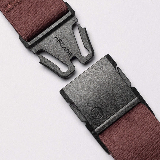 Close-up of a maroon stretch strap from Arcade Belt Co. by Arcade Belts, with a black metal buckle engraved ARCADE, set on a light surface. The low-profile buckle parts are aligned to snap together.