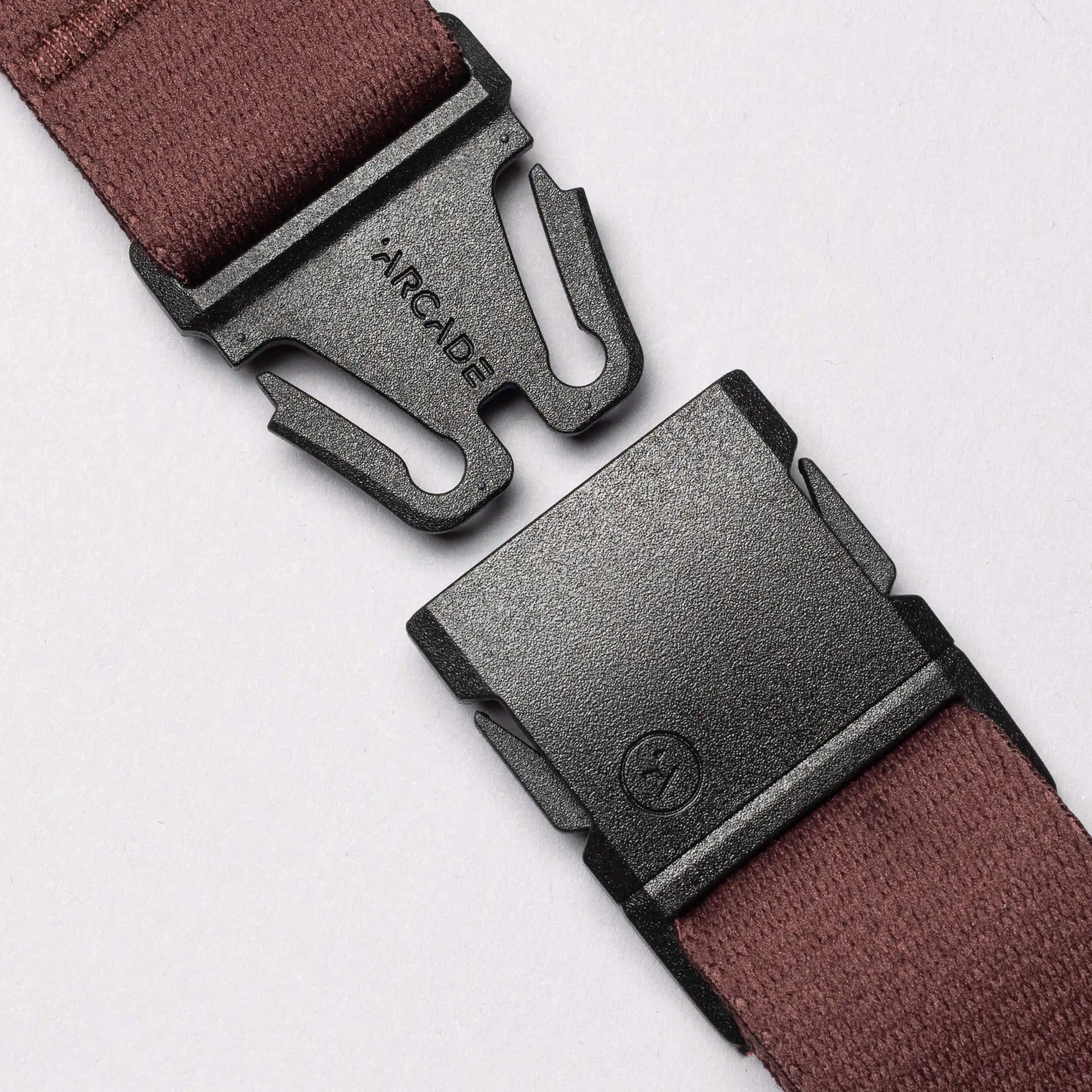 Close-up of a black plastic buckle on a maroon Arcade Belt Co. performance stretch belt by Arcade Belts, with "ARCADE" printed on one side, shown partially unclasped against a light background, displaying both connection points.