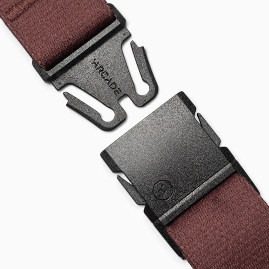 A close-up of a black plastic buckle, embossed with the Arcade Belts logo, is attached to a maroon Arcade Belt Co. performance stretch belt. The open buckle reveals its prongs and slot, while subtle pops of color from the strap add visual interest against a white background.