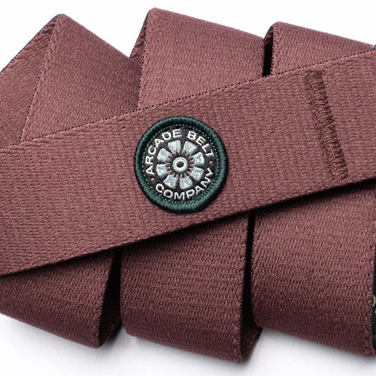 A close-up of a maroon performance stretch belt from Arcade Belt Co. by Arcade Belts, featuring textured material and a round black and white patch sewn on.