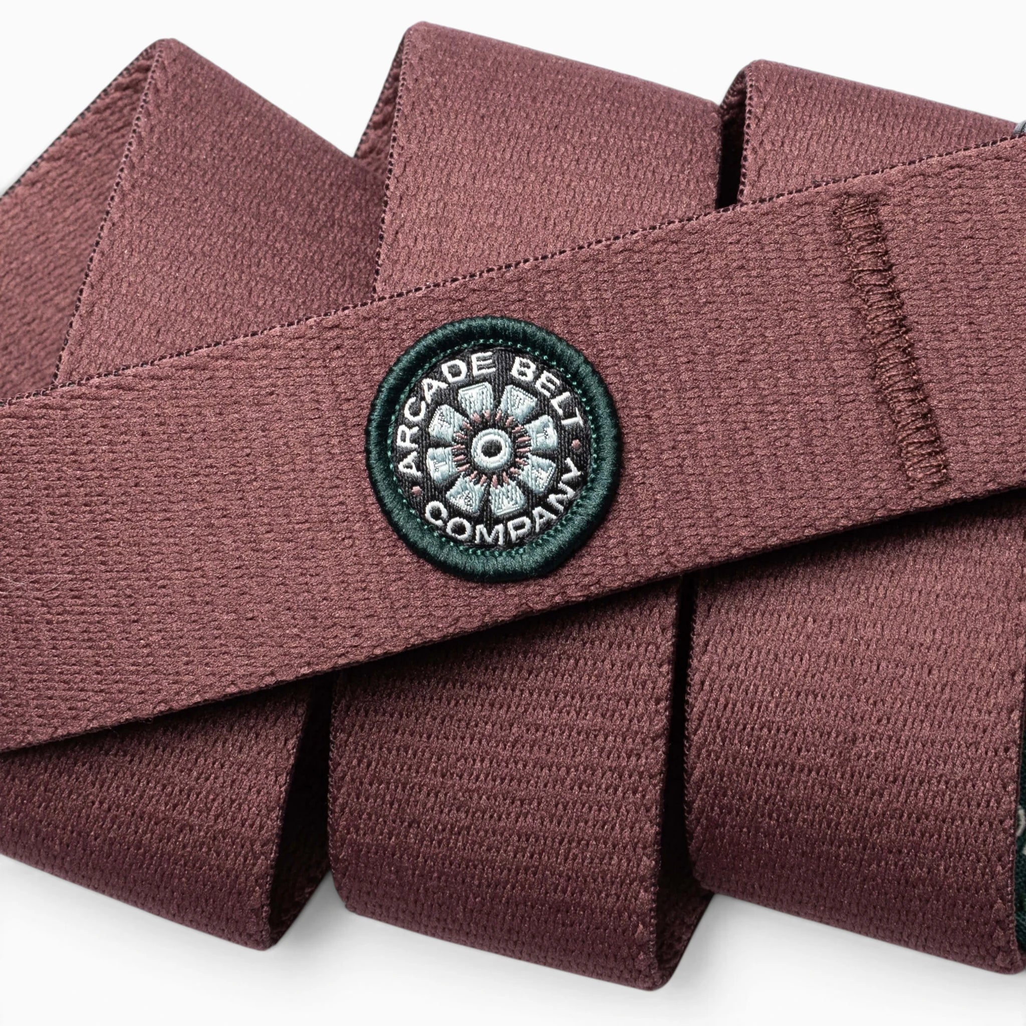 A close-up of a maroon Arcade Belt Co. performance stretch belt features a circular black patch with “Arcade Belts” in white text. This limited-edition patch design stands out against the folded layers, while subtle pops of color highlight its woven texture.