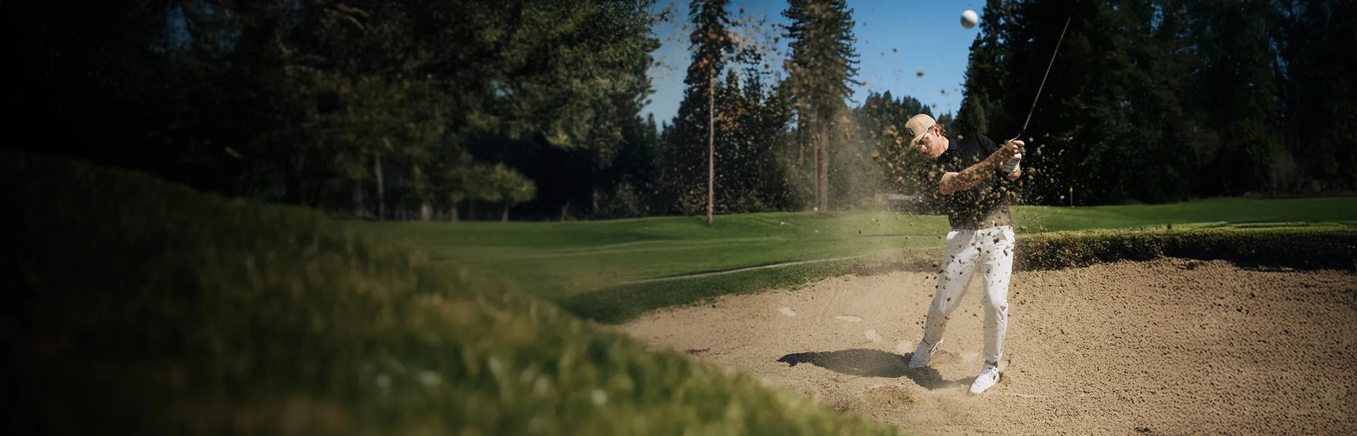 A golfer in a sand trap swings his club, sending sand and a golf ball into the air, with trees and green grass in the background on a sunny day.