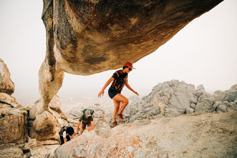 Three people hike among large, rugged rock formations. One steps up a boulder while two follow behind, all wearing hats, outdoor clothing, and practical Arcade Belts in a desert-like landscape.
