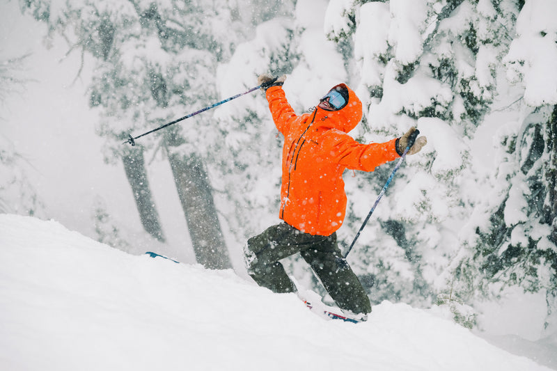 A skier wearing a bright orange jacket and goggles kneels in the snow with arms outstretched, surrounded by snow-covered trees during heavy snowfall.
