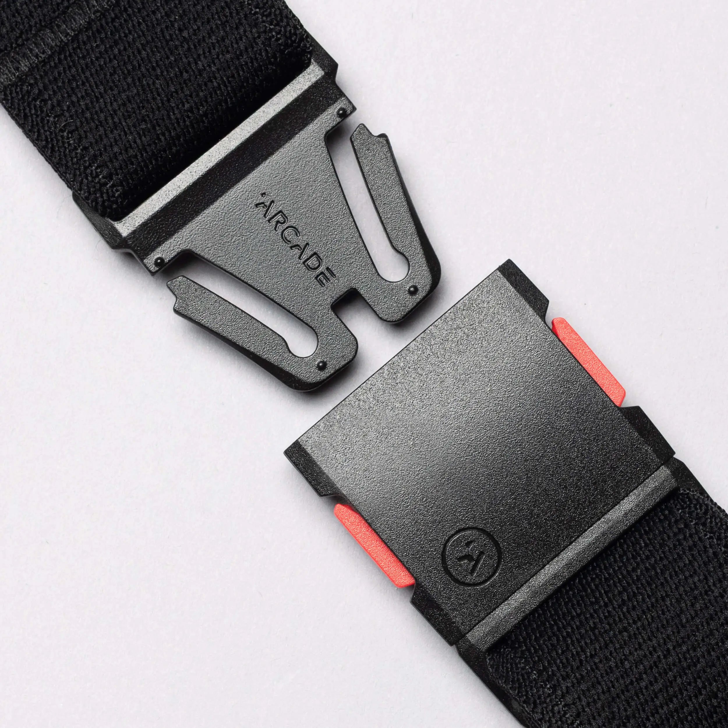 A close-up of the Arcade Belts Recco, a black performance stretch belt with a black metal buckle, partially unbuckled. The buckle displays "ARCADE" and has two red release buttons on its sides.