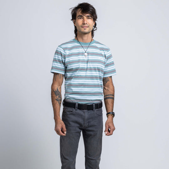 A man with dark hair, tattoos, and wearing a necklace and watch stands against a light gray background in a striped t-shirt, gray jeans, and the Midnight Kit belt by Arcade Belts.