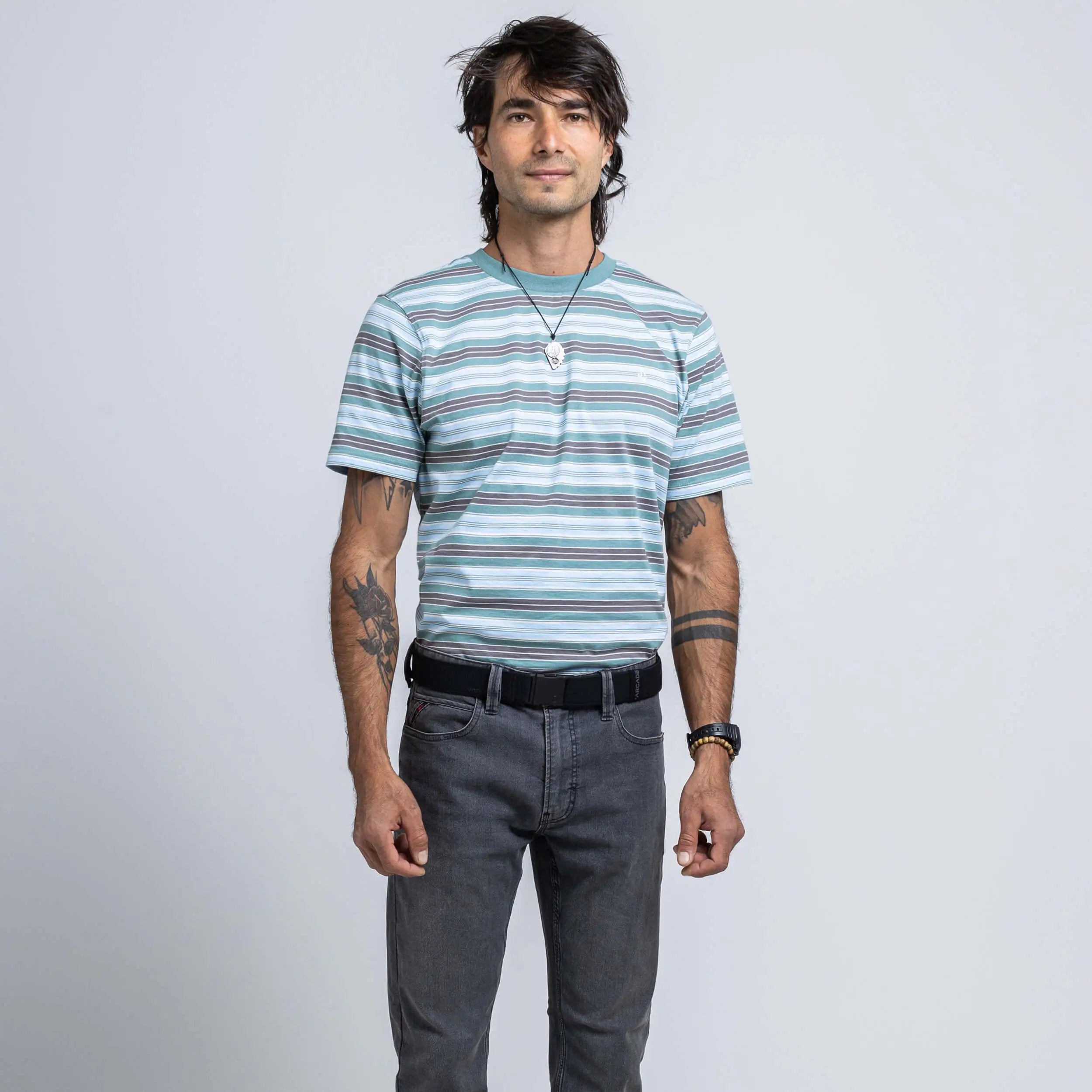 A person with dark hair, tattoos, and a necklace stands facing the camera against a plain light gray background, wearing a striped short-sleeve shirt and gray jeans, accessorized with the Atlas Gifting Kit from Arcade Belts that features the innovative A2 buckle system for enhanced performance stretch.