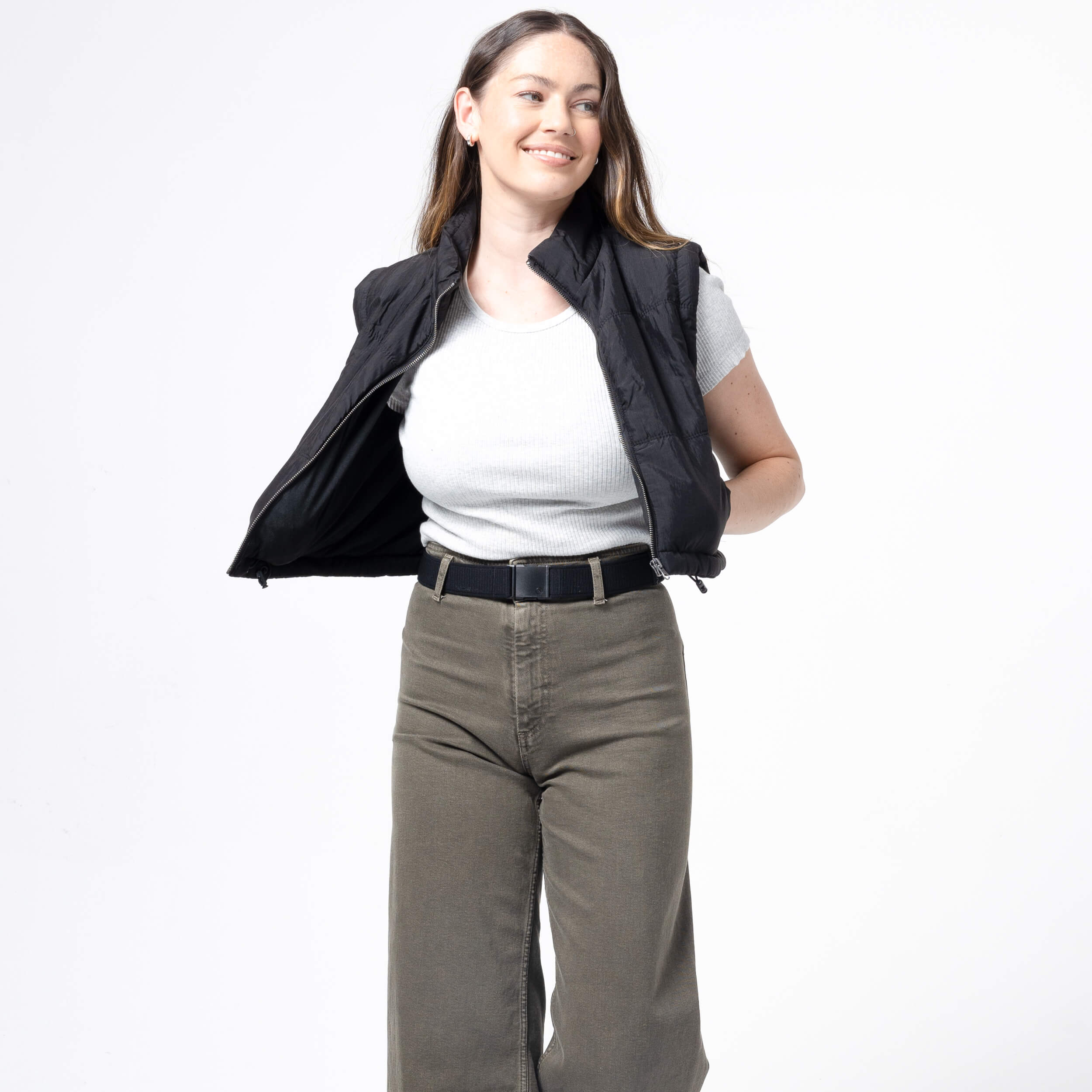 A woman with long brown hair, wearing the Atlas Gifting Kit by Arcade Belts—including a black vest over a light gray shirt and olive green performance stretch pants—smiles while posing with one arm behind her back against a white background.