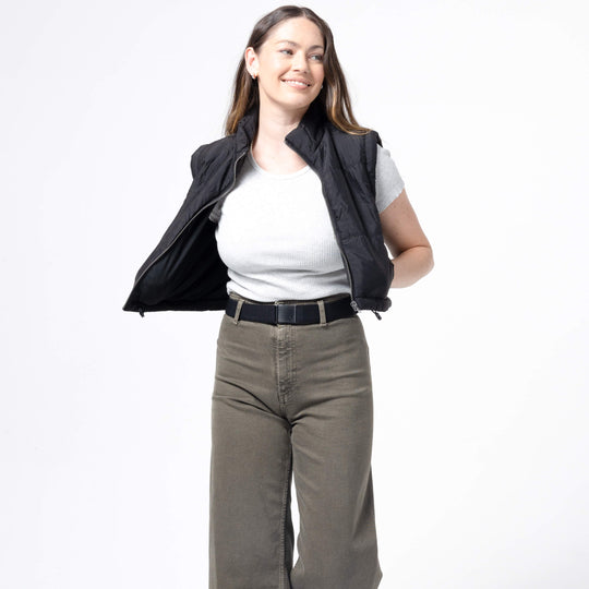 A woman with long brown hair, wearing the Atlas Gifting Kit by Arcade Belts—including a black vest over a light gray shirt and olive green performance stretch pants—smiles while posing with one arm behind her back against a white background.