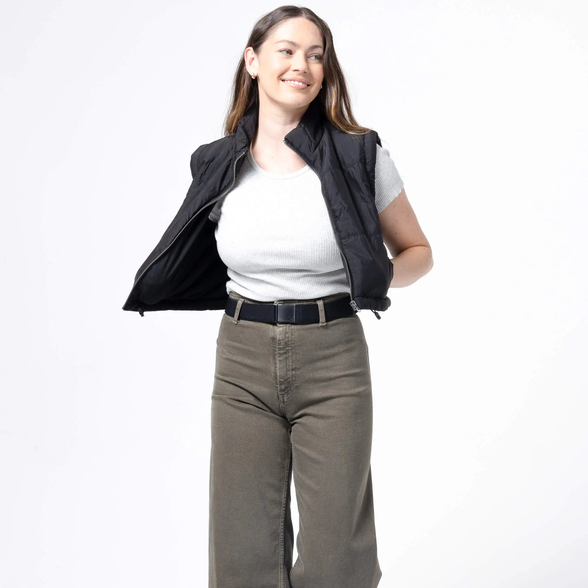 A woman with long brown hair, wearing the Atlas Gifting Kit by Arcade Belts—including a black vest over a light gray shirt and olive green performance stretch pants—smiles while posing with one arm behind her back against a white background.