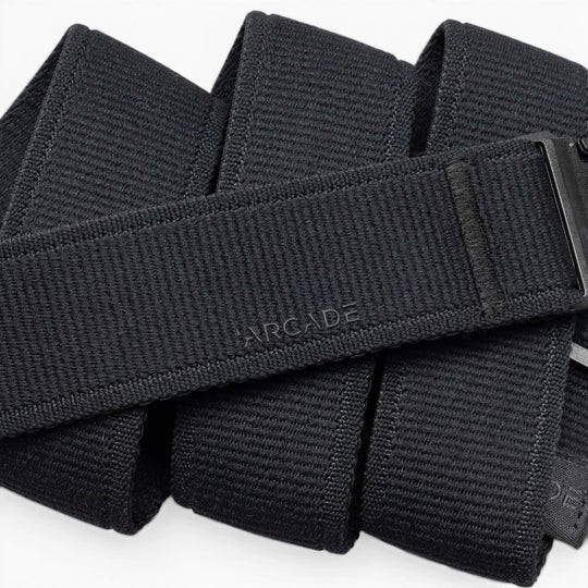 A close-up of the Atlas belt by Arcade Belts shows its black, textured design with ARCADE embossed on it. The belt is neatly folded, displaying a sleek plastic buckle partially visible on the right side.