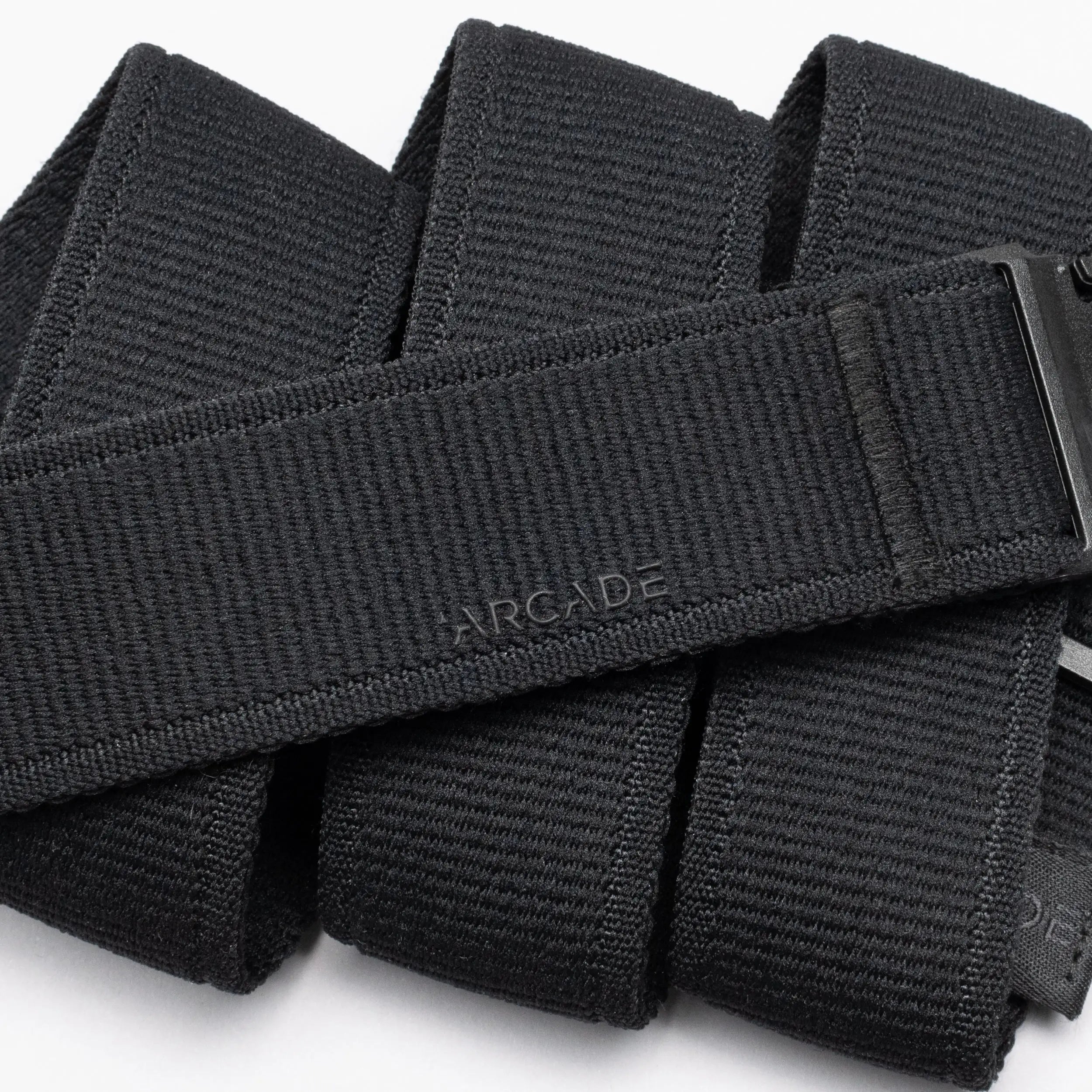 A close-up of the Atlas belt by Arcade Belts shows its black, textured design with ARCADE embossed on it. The belt is neatly folded, displaying a sleek plastic buckle partially visible on the right side.
