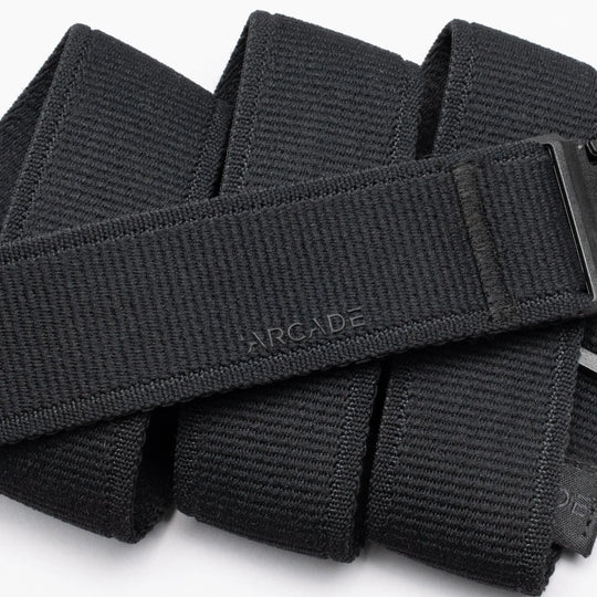 A close-up of the Atlas belt by Arcade Belts shows its black, textured design with ARCADE embossed on it. The belt is neatly folded, displaying a sleek plastic buckle partially visible on the right side.
