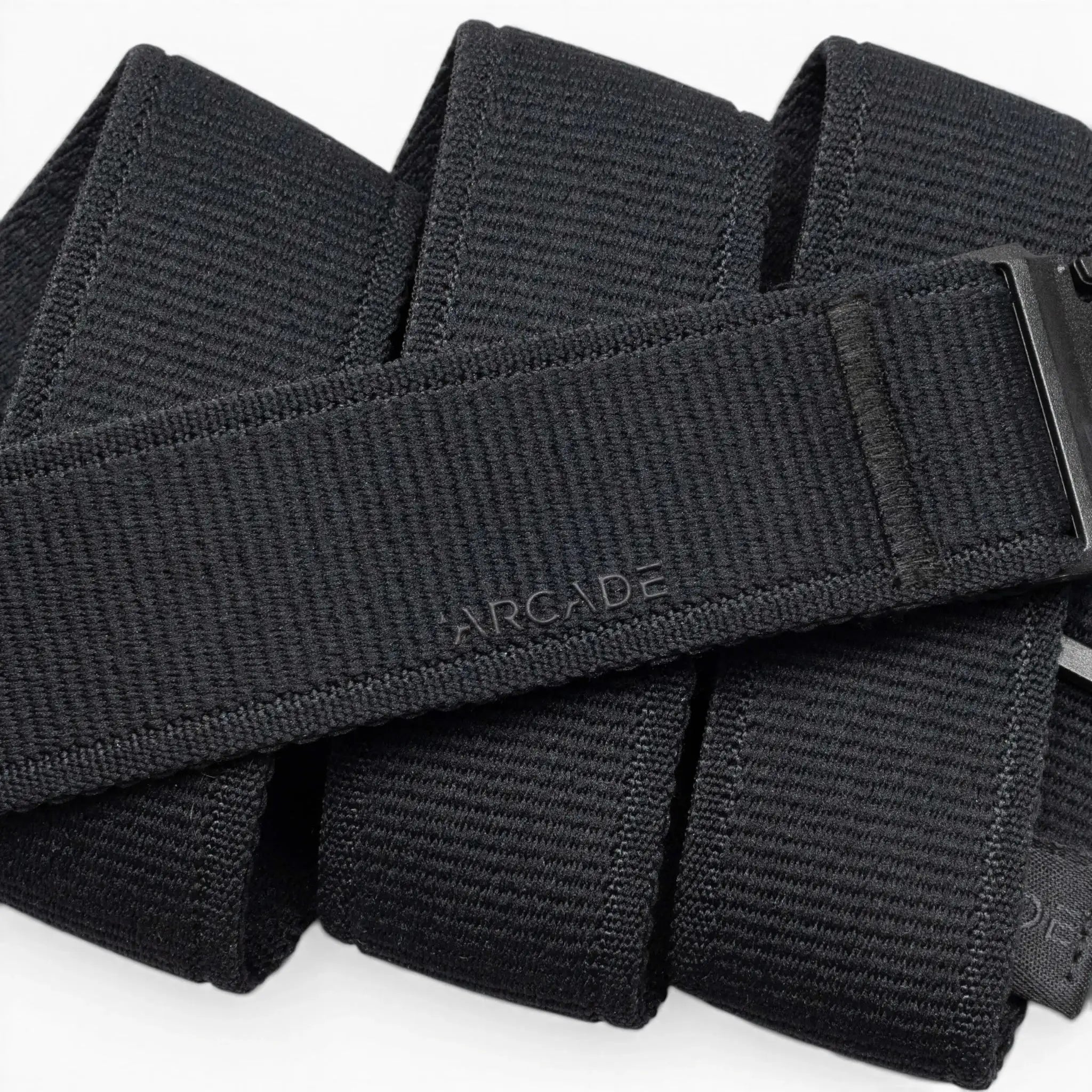A close-up of the Atlas belt by Arcade Belts shows its black, textured design with ARCADE embossed on it. The belt is neatly folded, displaying a sleek plastic buckle partially visible on the right side.