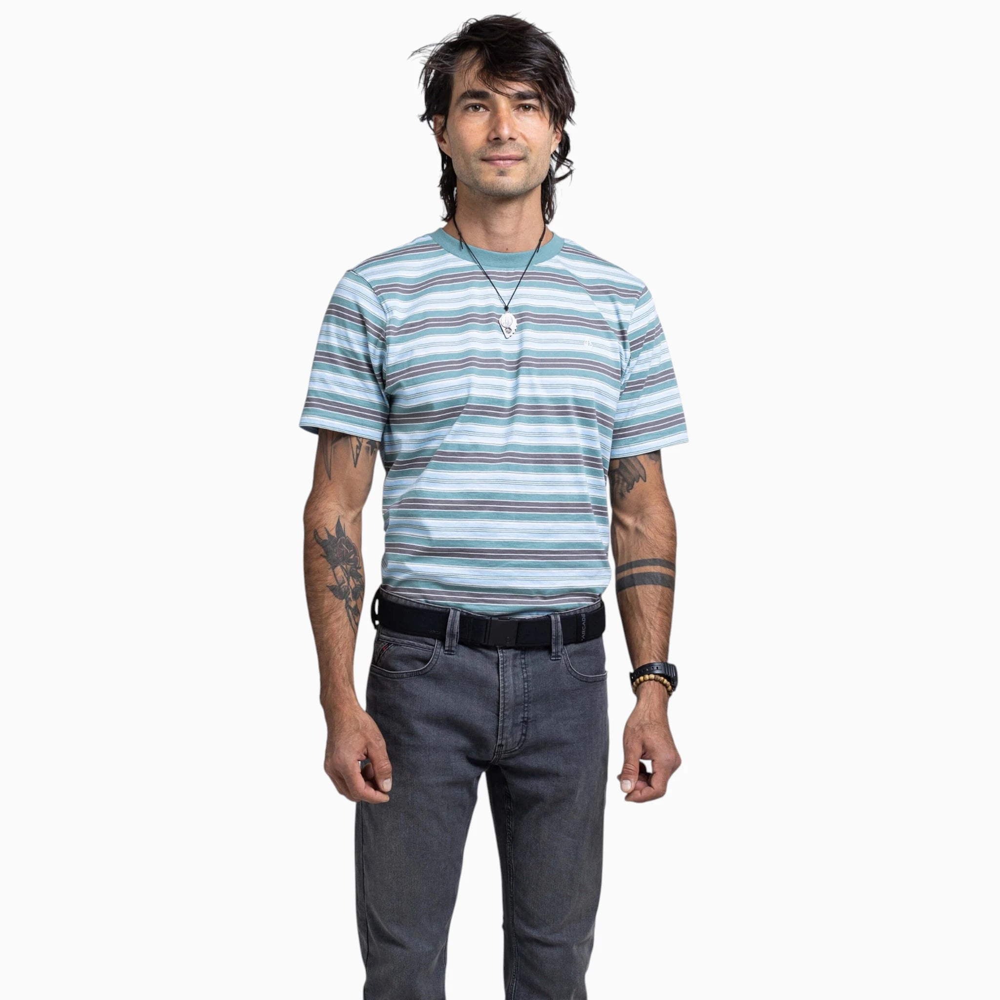 A person with dark hair, tattoos on both arms, a necklace, and a wristwatch stands facing forward against a plain white background. They are wearing a striped T-shirt, dark jeans, and the Atlas Gifting Kit by Arcade Belts, featuring the signature A2 buckle system.
