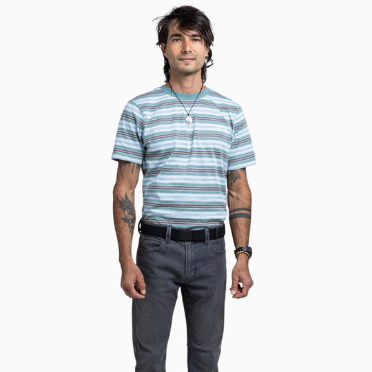 A person with dark hair, tattoos on both arms, a necklace, and a wristwatch stands facing forward against a plain white background. They are wearing a striped T-shirt, dark jeans, and the Atlas Gifting Kit by Arcade Belts, featuring the signature A2 buckle system.