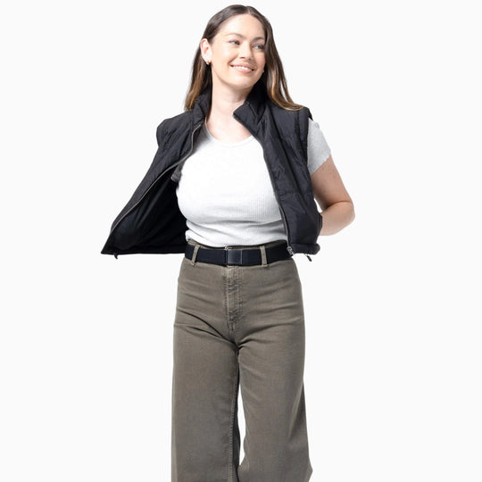 A woman with long brown hair smiles while wearing the Atlas Gifting Kit by Arcade Belts, which includes a gray t-shirt, olive green performance stretch pants for comfort, and a partially unzipped black vest. She stands against a plain white background.