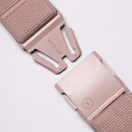A close-up of the pink Atlas Stretch Webbing belt by Arcade Belts, featuring a plastic buckle, ARCADE embossed on one side, and a small smiley face icon on the other, set against a plain light pink background.