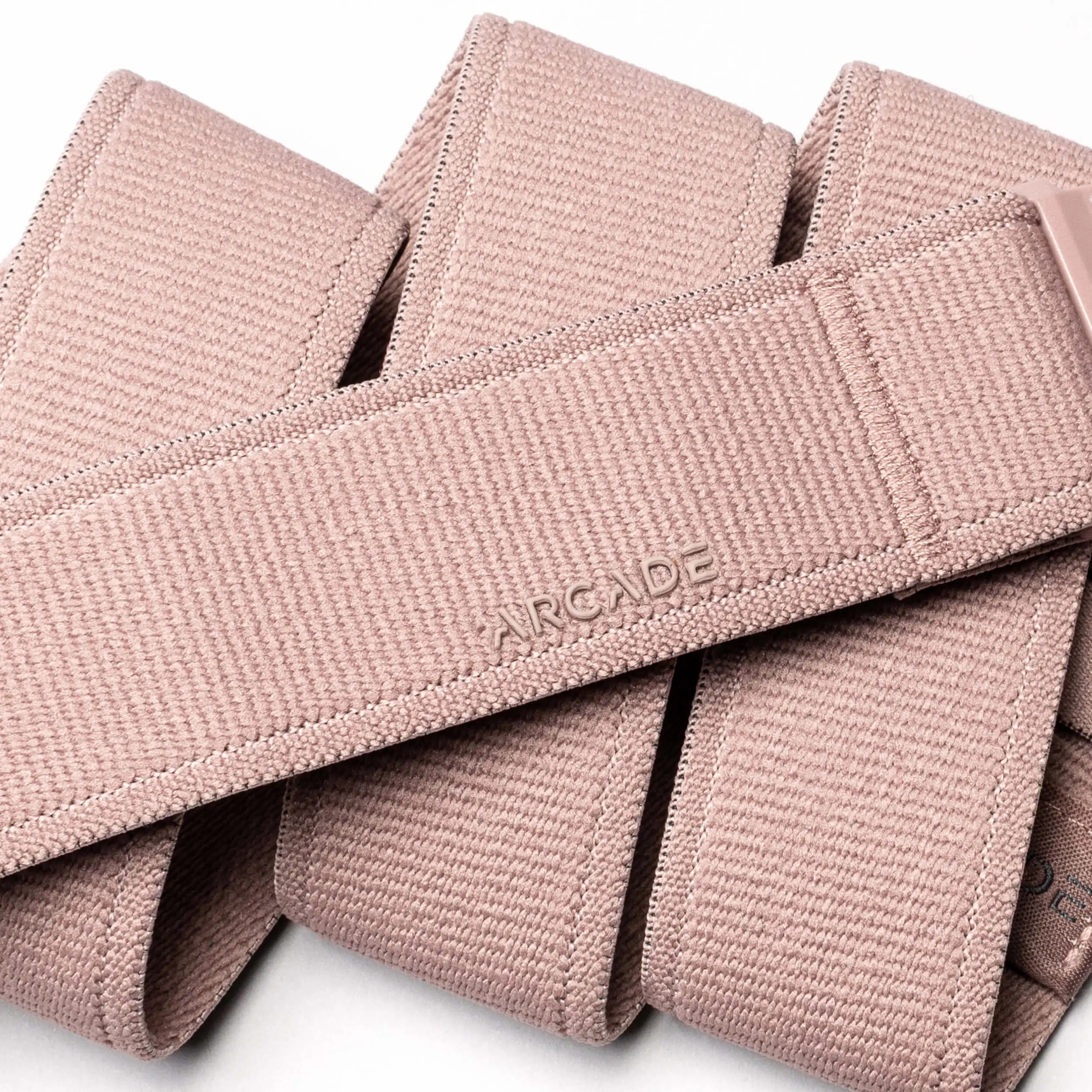 A close-up of the Atlas belt by Arcade Belts, featuring light pink elastic stretch material, ARCADE embossed branding, visible stitching details, textured finish, and a sleek low-profile design.
