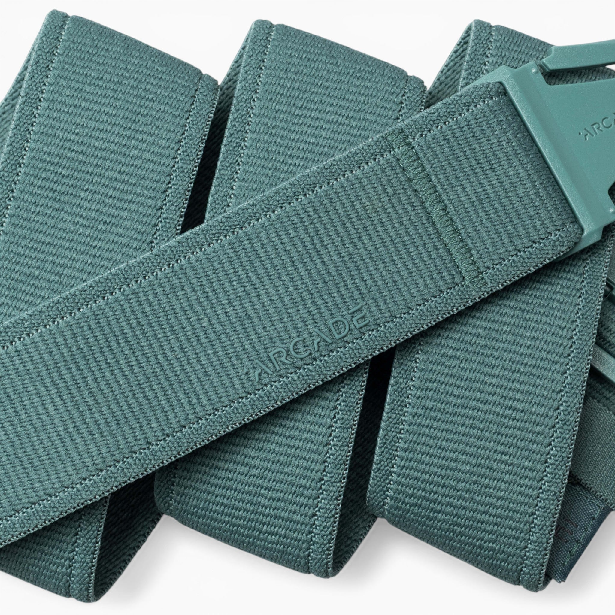 A close-up of the teal woven Atlas belt by Arcade Belts, featuring a matching plastic A2 buckle. The belt is folded over itself, with the word “ARCADE” subtly embossed on its surface, highlighting the innovative design of performance stretch belts from Arcade Belts’ Atlas collection.