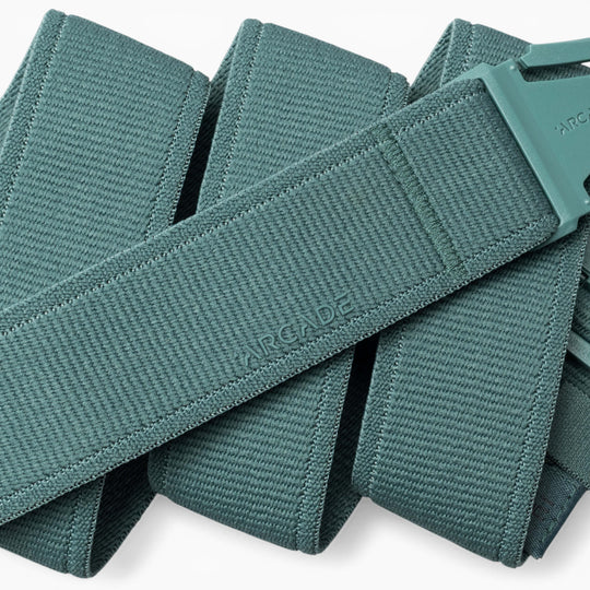 A close-up of the teal woven Atlas belt by Arcade Belts, featuring a matching plastic A2 buckle. The belt is folded over itself, with the word “ARCADE” subtly embossed on its surface, highlighting the innovative design of performance stretch belts from Arcade Belts’ Atlas collection.