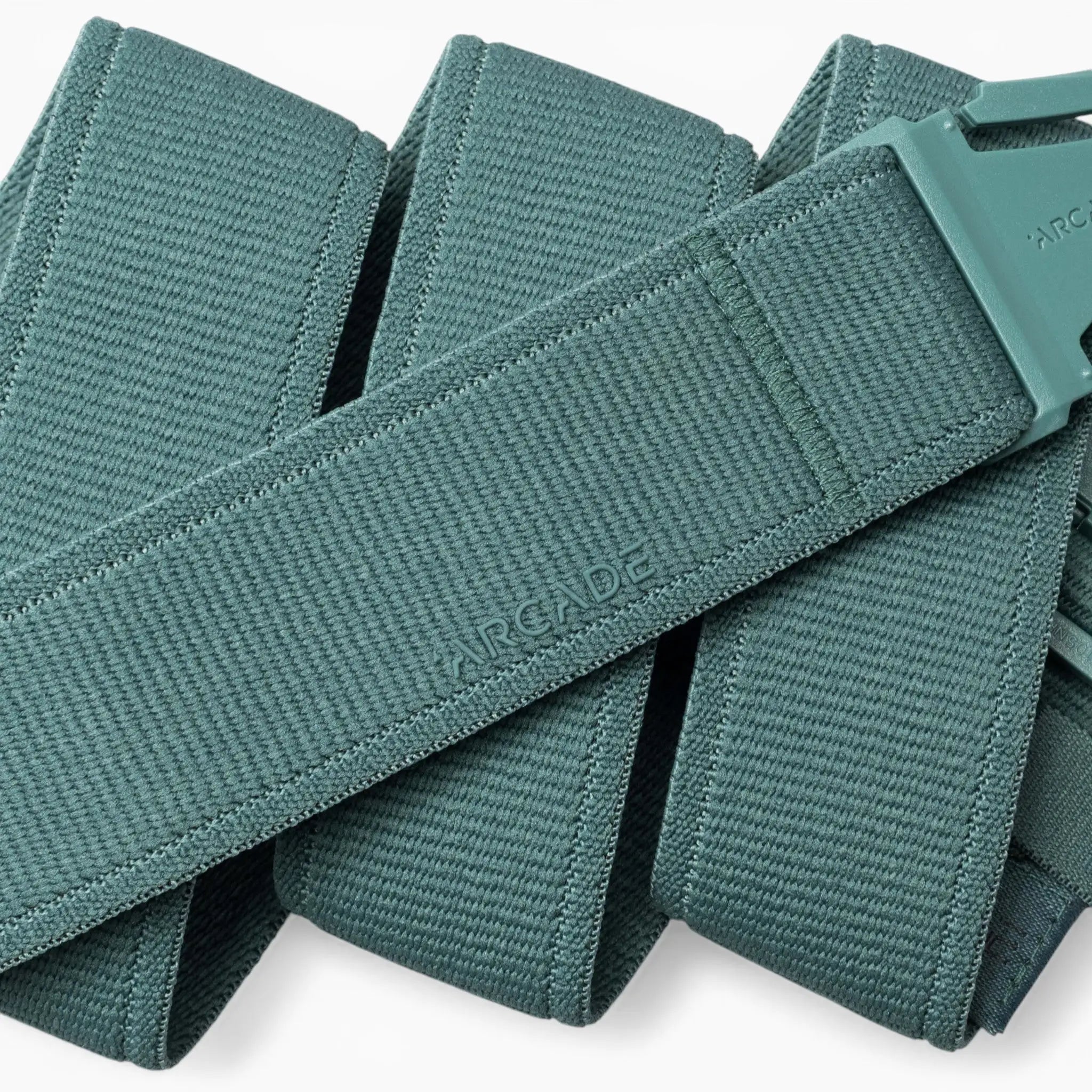 A close-up of the teal woven Atlas belt by Arcade Belts, featuring a matching plastic A2 buckle. The belt is folded over itself, with the word “ARCADE” subtly embossed on its surface, highlighting the innovative design of performance stretch belts from Arcade Belts’ Atlas collection.