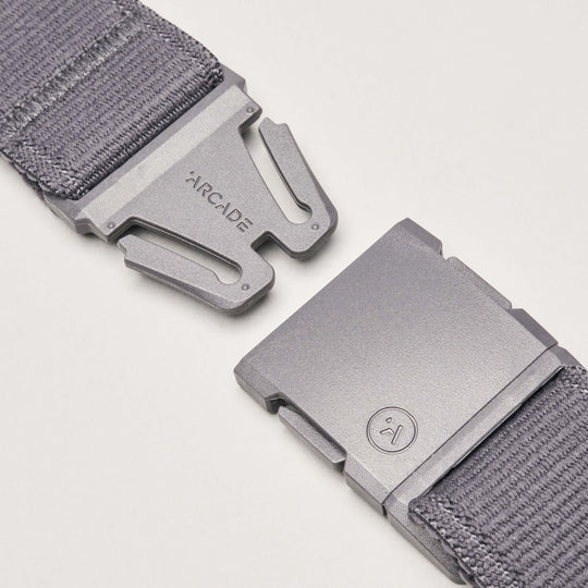 Close-up of the Atlas belt by Arcade Belts in gray stretch webbing, featuring a metal buckle. One side is engraved with the brand name; the other shows a small circular smiley face logo. Buckle pieces are set apart on a white background.