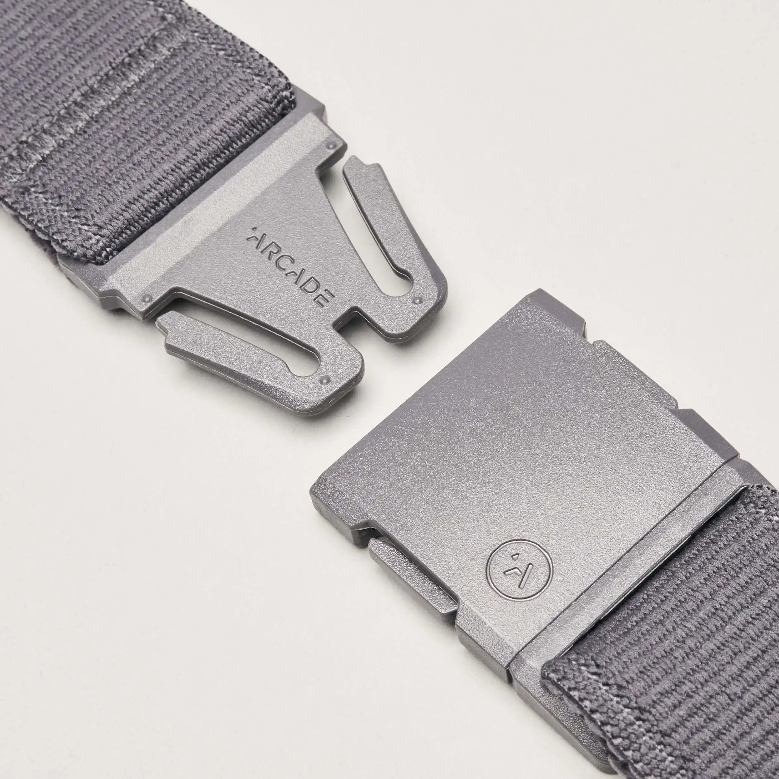 Close-up of the Atlas belt by Arcade Belts in gray stretch webbing, featuring a metal buckle. One side is engraved with the brand name; the other shows a small circular smiley face logo. Buckle pieces are set apart on a white background.
