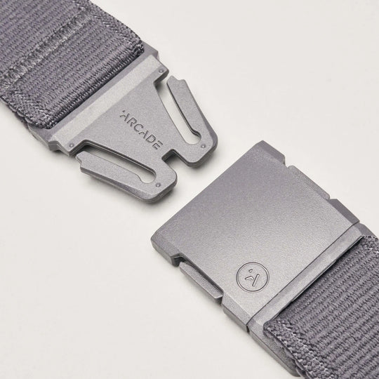 Close-up of the Atlas belt by Arcade Belts in gray stretch webbing, featuring a metal buckle. One side is engraved with the brand name; the other shows a small circular smiley face logo. Buckle pieces are set apart on a white background.