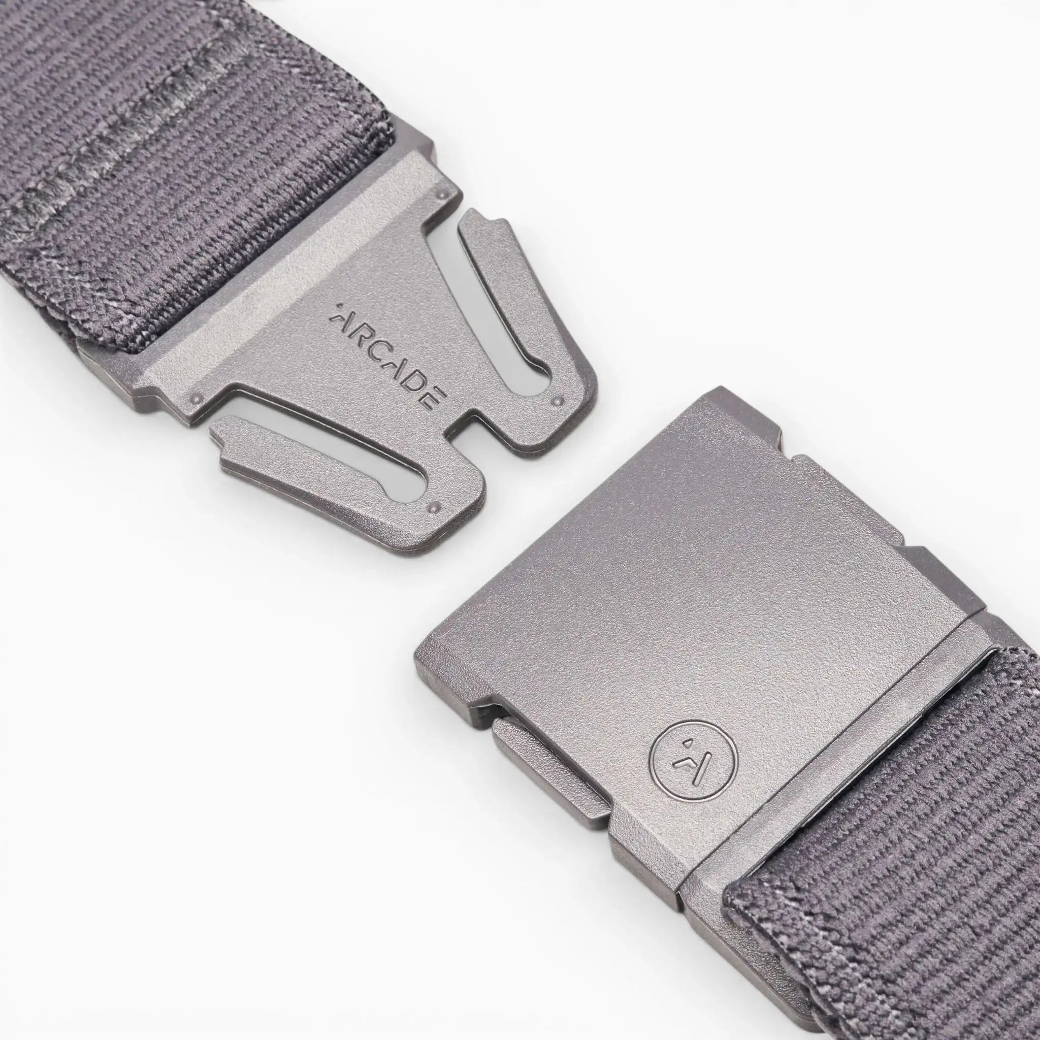 Close-up of the Atlas belt by Arcade Belts in gray stretch webbing, featuring a metal buckle. One side is engraved with the brand name; the other shows a small circular smiley face logo. Buckle pieces are set apart on a white background.
