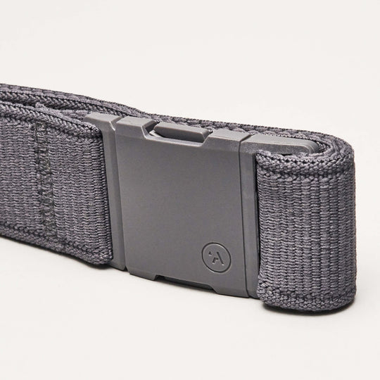 A close-up of an Arcade Belts Atlas dark gray woven Elastic Stretch Belt with a rectangular matte gray, low-profile buckle featuring a circular logo with an upward-pointing arrow, resting on a light background.