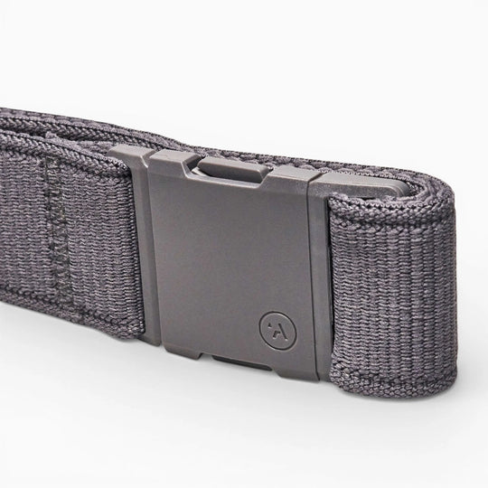 A close-up of an Arcade Belts Atlas dark gray woven Elastic Stretch Belt with a rectangular matte gray, low-profile buckle featuring a circular logo with an upward-pointing arrow, resting on a light background.