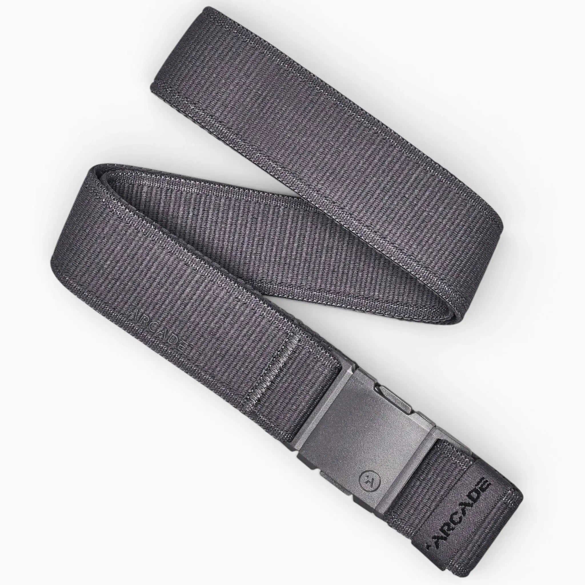 The Atlas by Arcade Belts is a dark gray low-profile belt with a rectangular plastic buckle, shown coiled on a white background. ARCADE is printed near the buckle, making it perfect for travel or daily use.