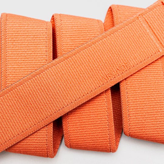 Four neatly folded orange Atlas straps from Arcade Belts, crafted from stretch webbing, are arranged in parallel; one diagonal shows the subtly embossed ARCADE logo—perfect as travel belts.