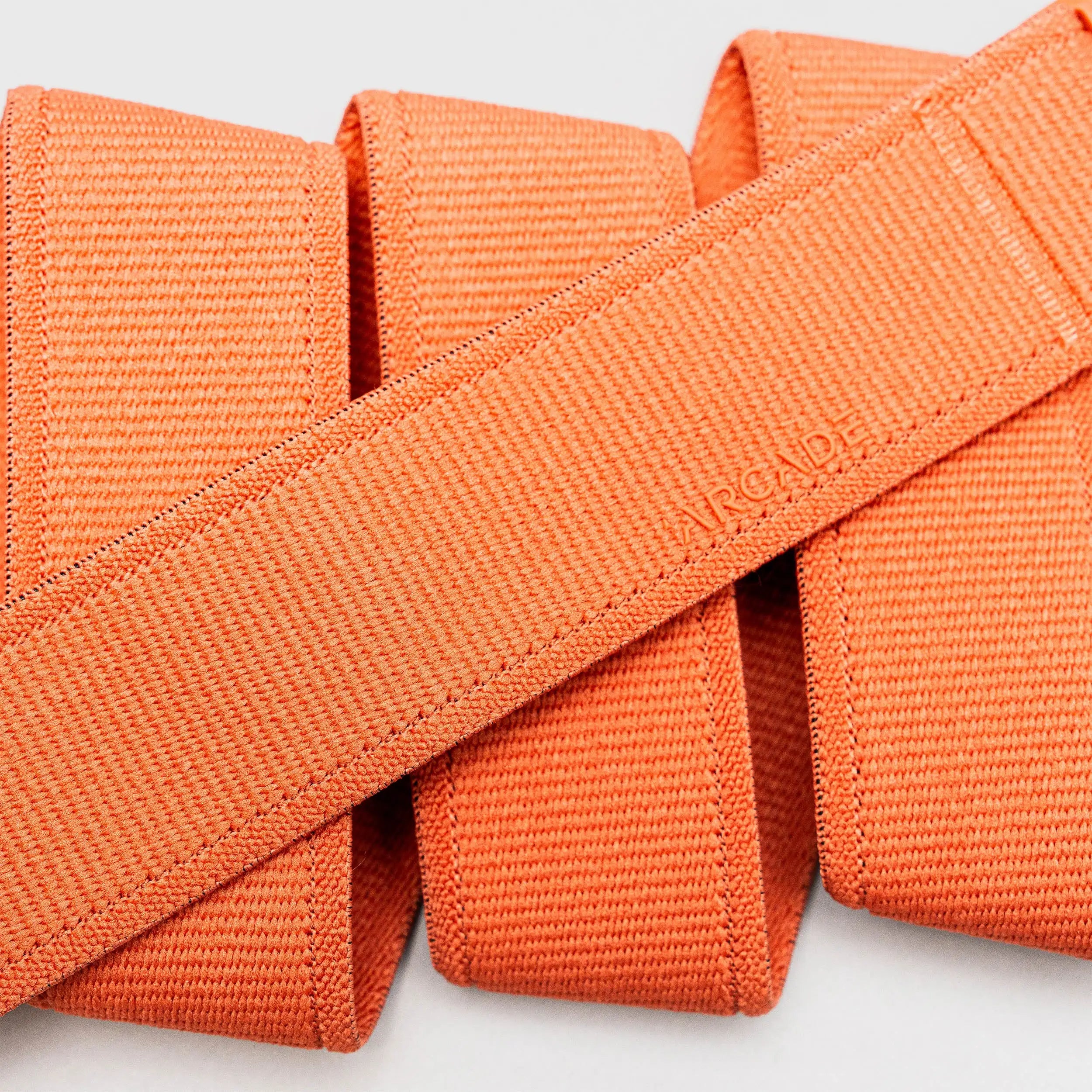 Four neatly folded orange Atlas straps from Arcade Belts, crafted from stretch webbing, are arranged in parallel; one diagonal shows the subtly embossed ARCADE logo—perfect as travel belts.