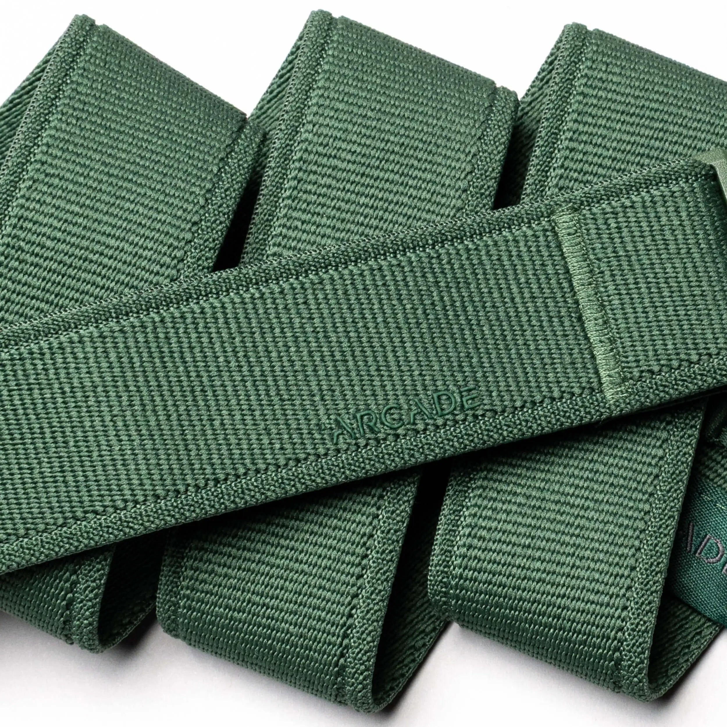 A close-up of green woven straps from the Atlas belt by Arcade Belts, featuring a plastic buckle and the word ARCADE embossed on one strap, arranged in an overlapping pattern on a white background.
