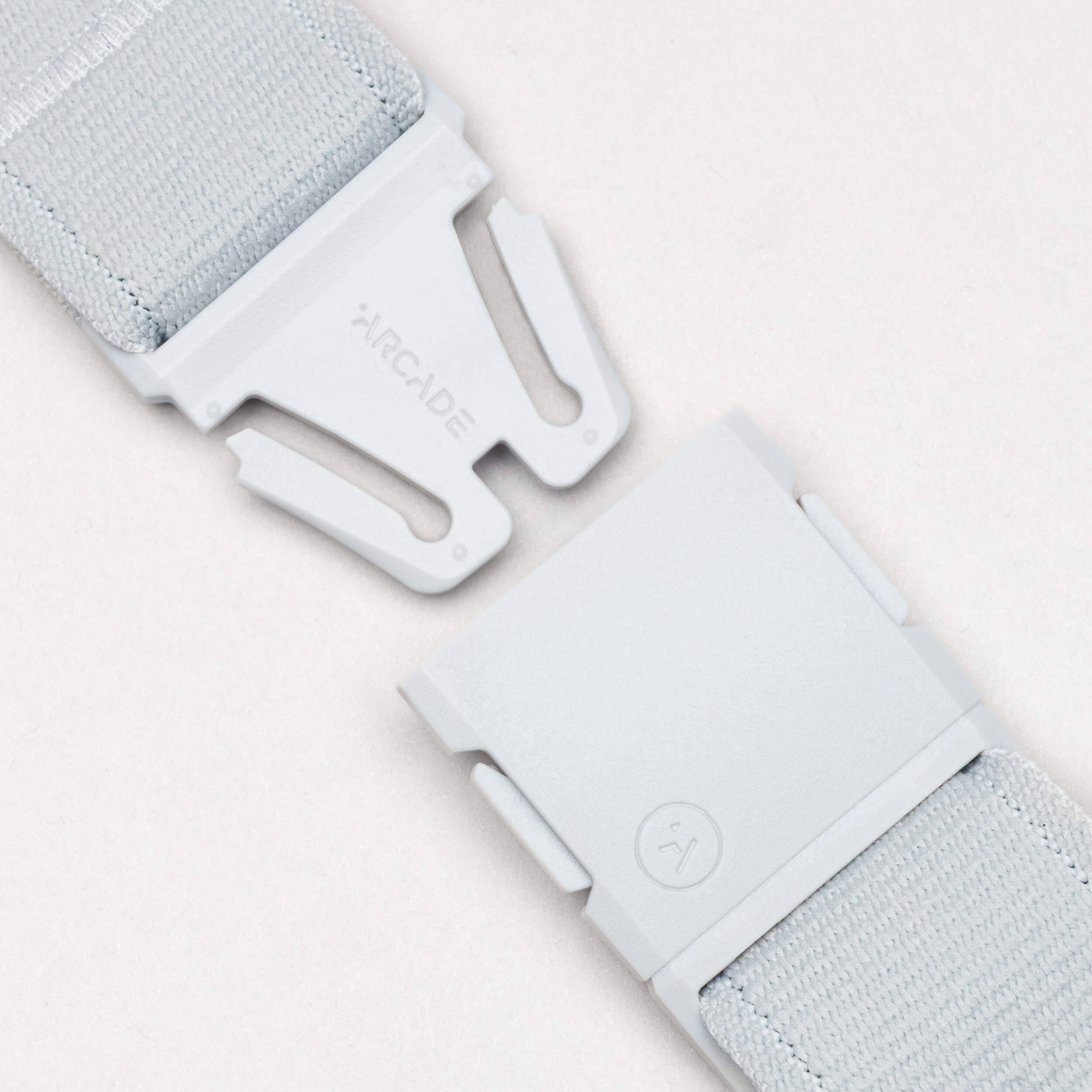 A close-up of the Atlas belt by Arcade Belts in light gray, featuring a matching plastic buckle—partially unclasped—with "ARCADE" on one side and a small logo on the other, showcasing its comfortable performance stretch design.