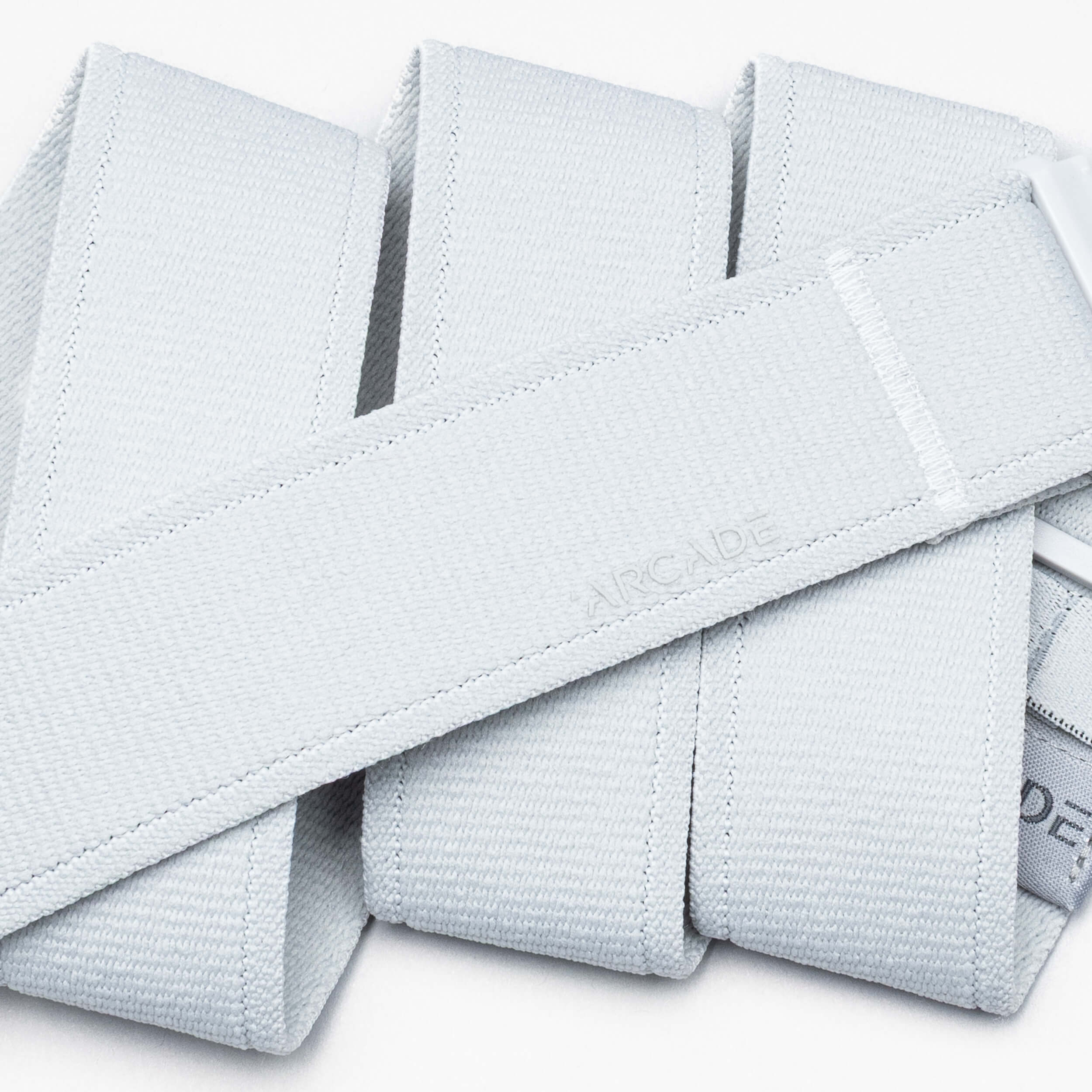 A close-up of the light gray Atlas performance stretch belt by Arcade Belts, featuring an A2 buckle and a subtle ARCADE emboss, folded in an overlapping pattern on a white background.