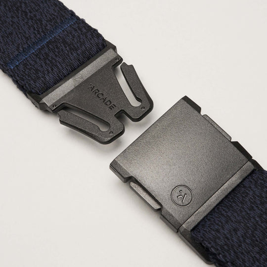 Close-up of a black metal buckle on the navy blue Atlas belt by Arcade Belts, partially unclasped with ARCADE engraved on the clasp, displayed against a plain white background.