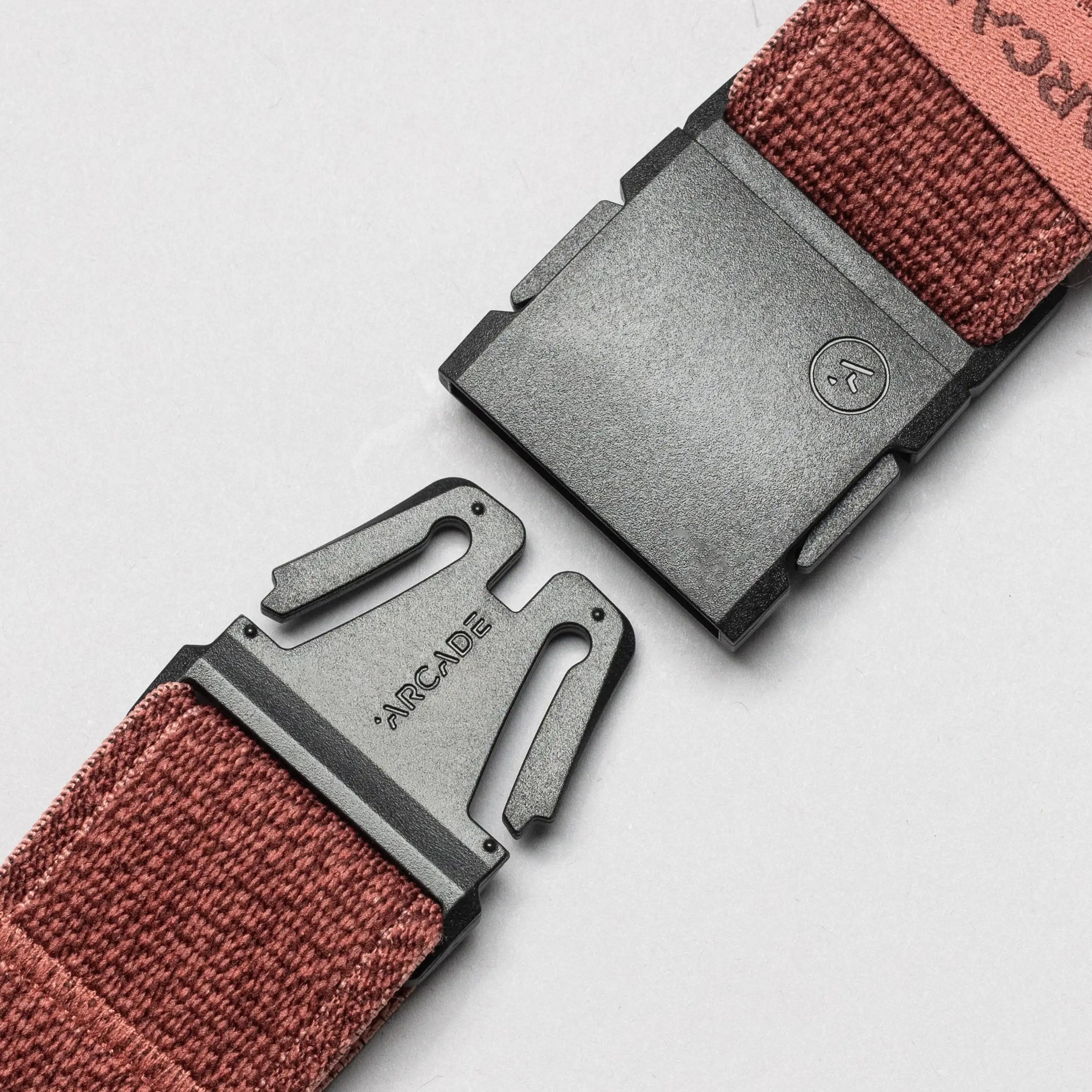 Close-up of the maroon Atlas Low Profile Belt by Arcade Belts, crafted from stretch webbing with a black plastic buckle. ARCADE is engraved on the metal clasp, ready to fasten. The background is plain light gray.