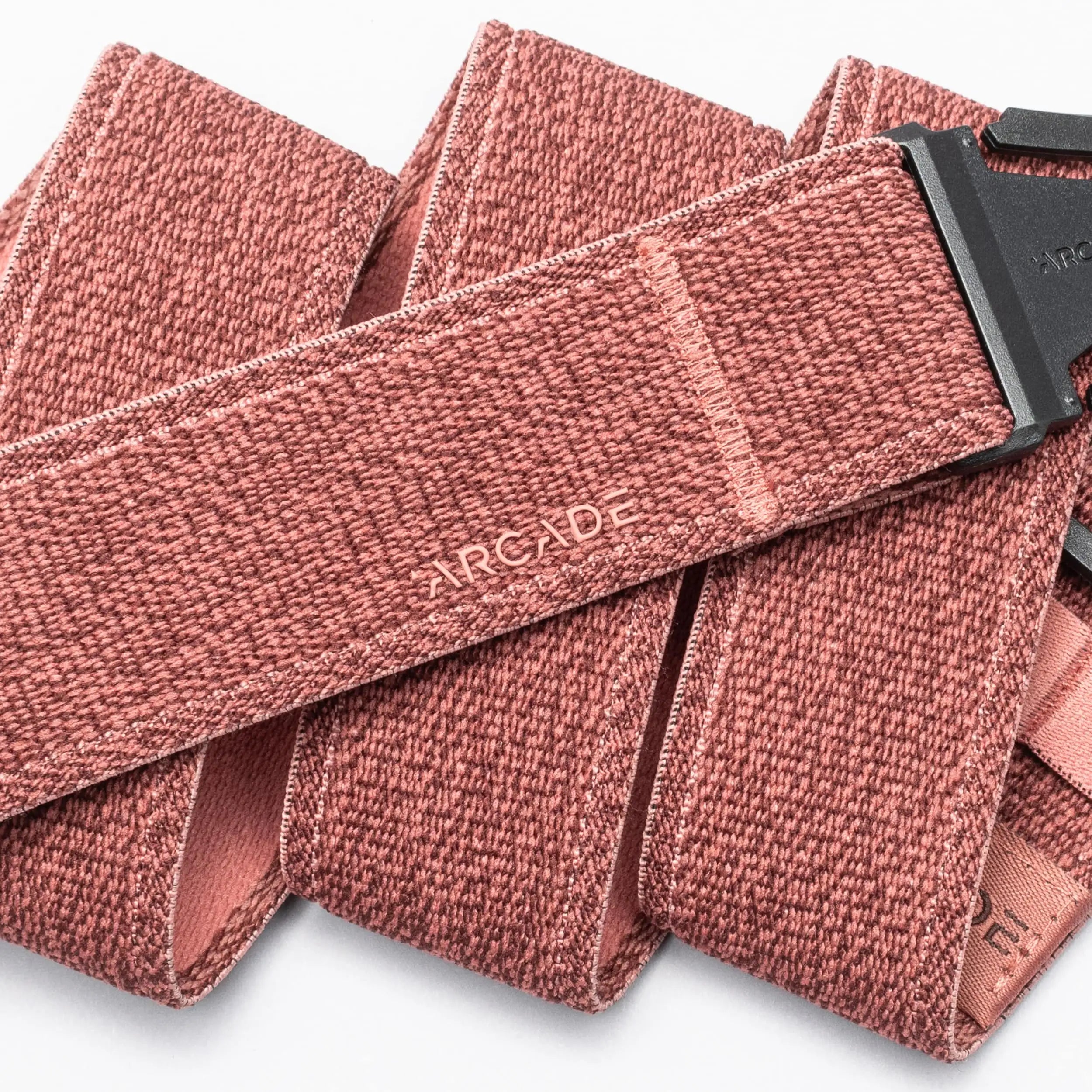 A close-up of the Atlas belt by Arcade Belts shows its reddish-pink textured elastic with ARCADE stitched in white and a low-profile plastic buckle, laid out in overlapping folds.