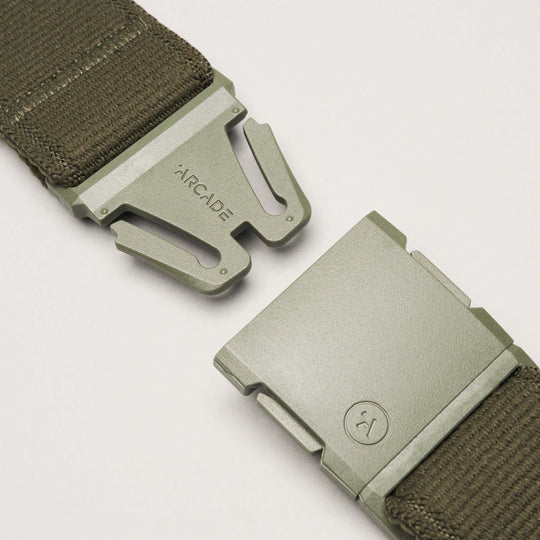 Close-up of an olive green Atlas belt from Arcade Belts with a low-profile metal buckle, partially unclasped. ARCADE is engraved on one side and a circular logo appears on the other. The belt is crafted from durable fabric.
