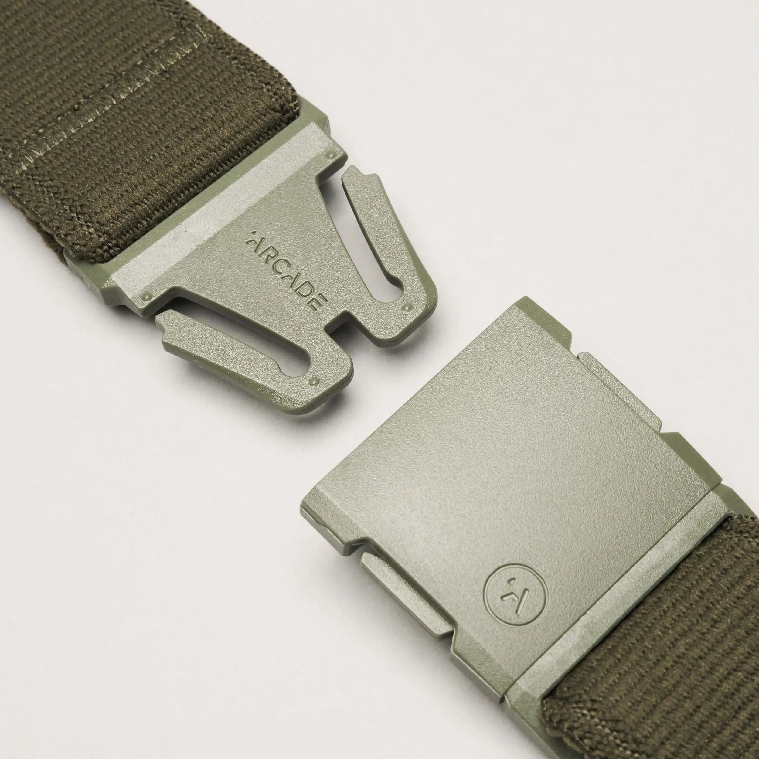 Close-up of an olive green Atlas belt from Arcade Belts with a low-profile metal buckle, partially unclasped. ARCADE is engraved on one side and a circular logo appears on the other. The belt is crafted from durable fabric.
