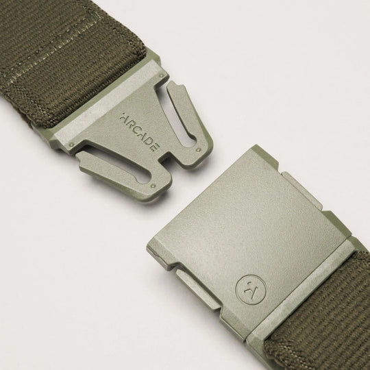 Close-up of an olive green Atlas belt from Arcade Belts with a low-profile metal buckle, partially unclasped. ARCADE is engraved on one side and a circular logo appears on the other. The belt is crafted from durable fabric.