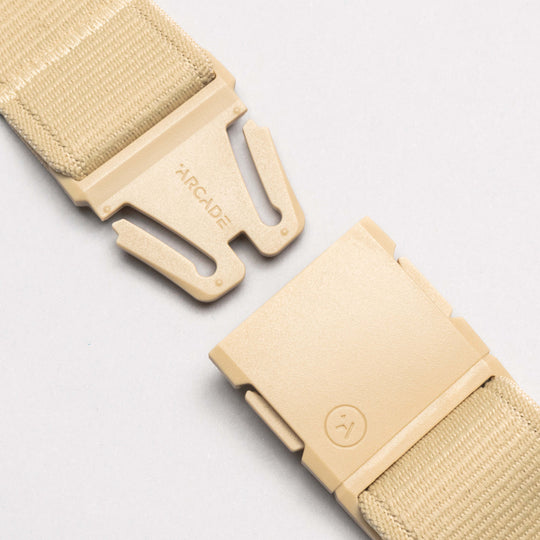 Close-up of the Atlas tan adjustable strap by Arcade Belts, featuring stretch webbing and a plastic buckle. The separated buckle shows the Arcade Belts logo and branding, all set against a light gray background.