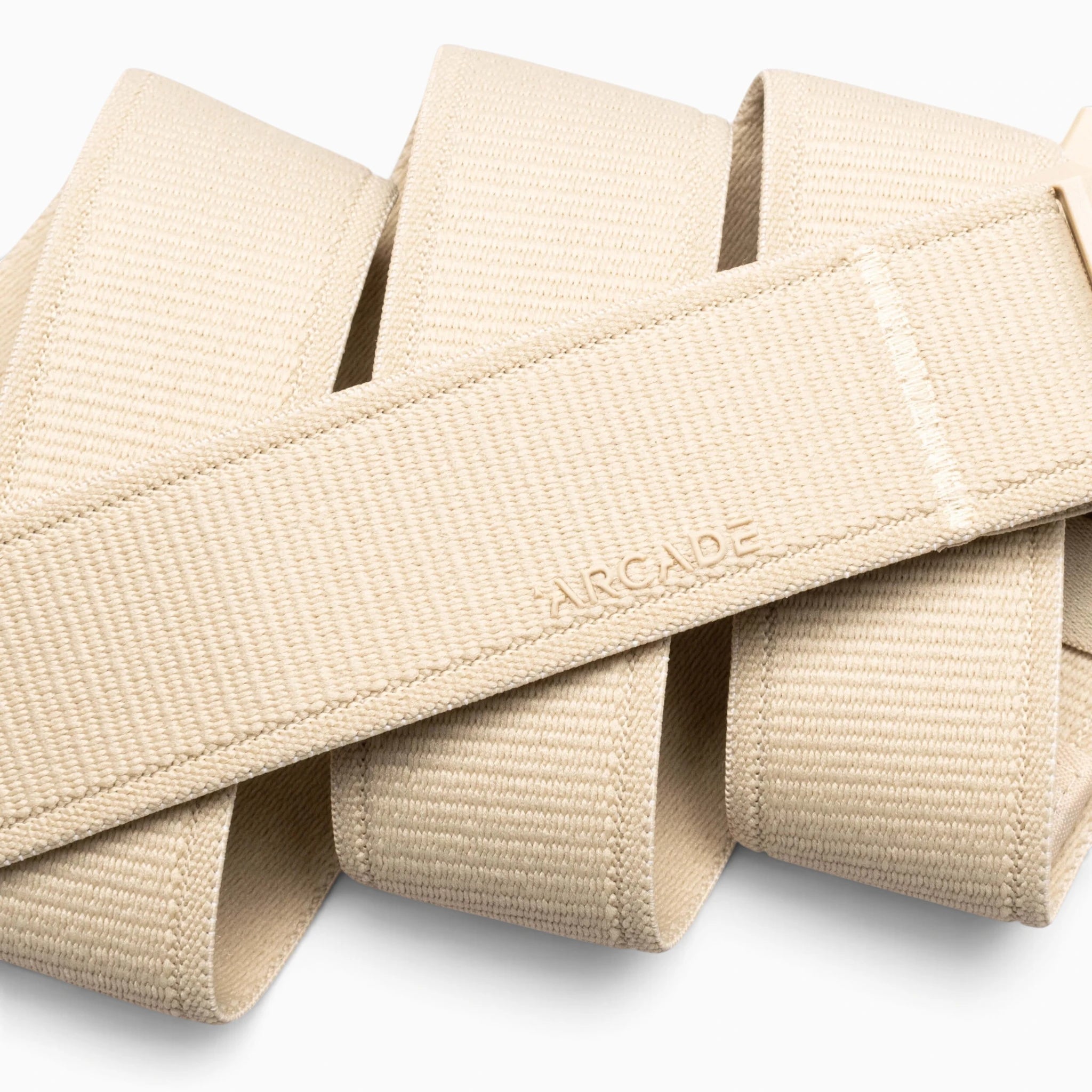 A close-up of the Atlas adventure belt by Arcade Belts, featuring beige woven fabric embossed with “ARCADE.” The performance stretch belt is folded in wide, even loops against a white background.